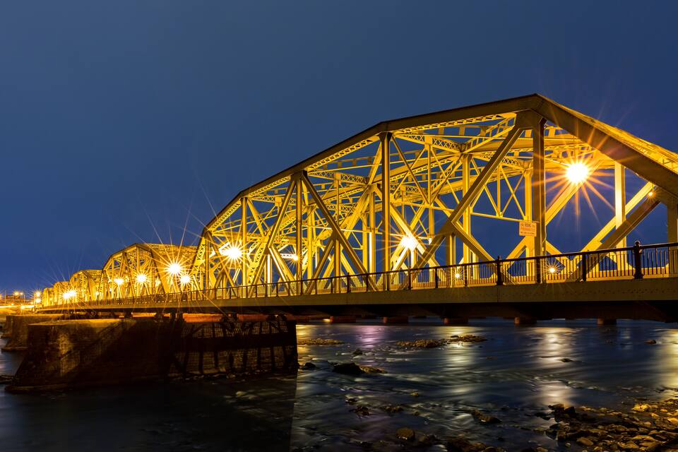 A steel bridge at night with lights reflecting on the water below.