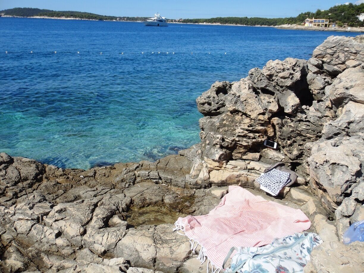 A rocky shoreline with clear blue water, a yacht in the distance, and a beach house on the horizon.