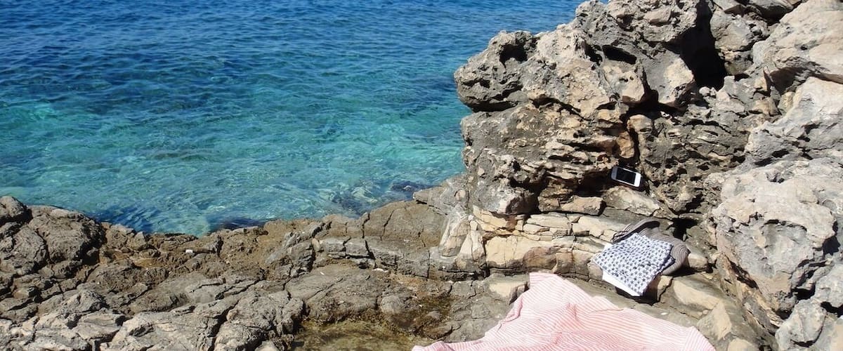 A rocky shoreline with clear blue water, a yacht in the distance, and a beach house on the horizon.