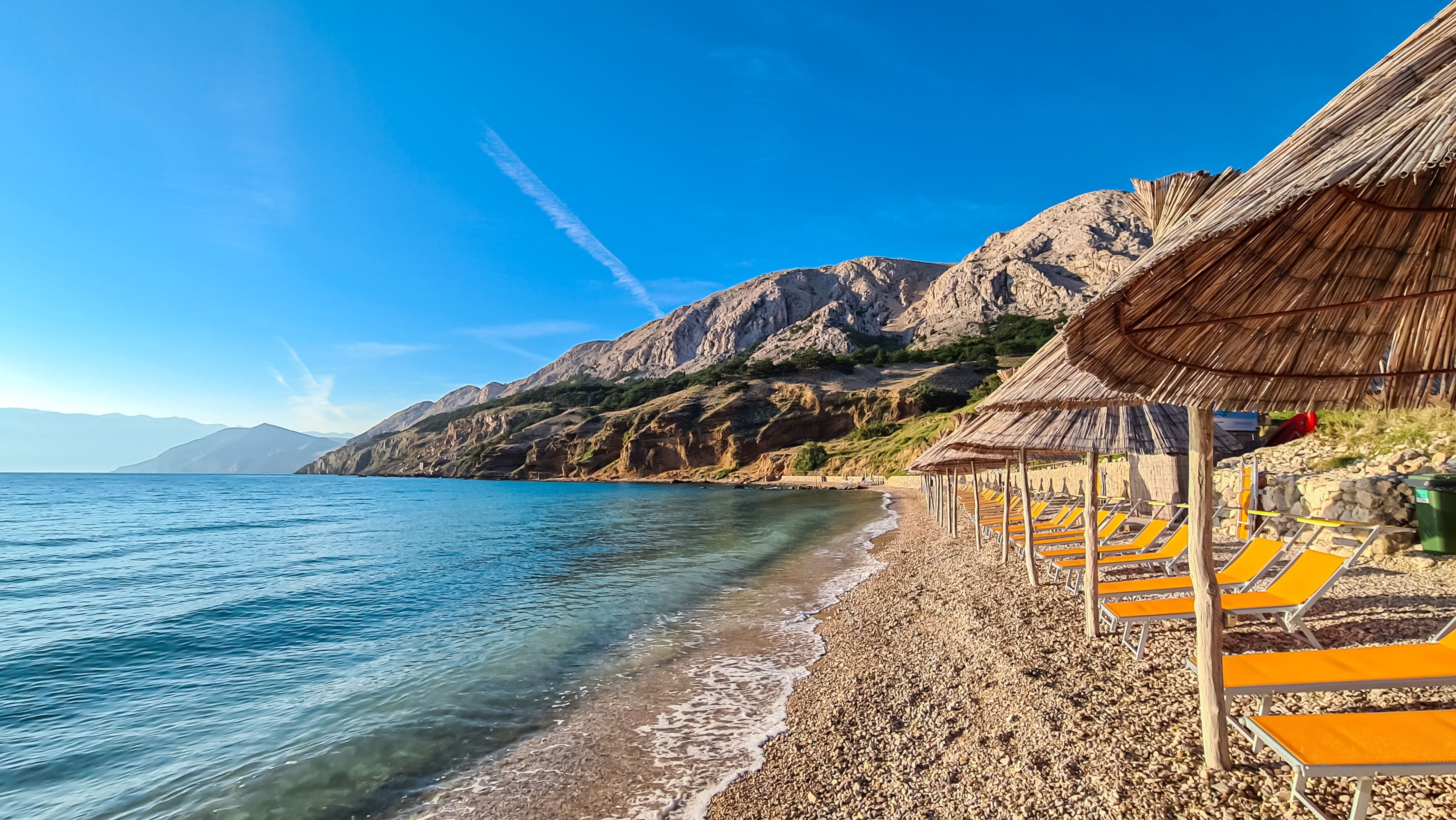 Una playa de guijarros con sombrillas de paja y una imponente montaña al fondo.