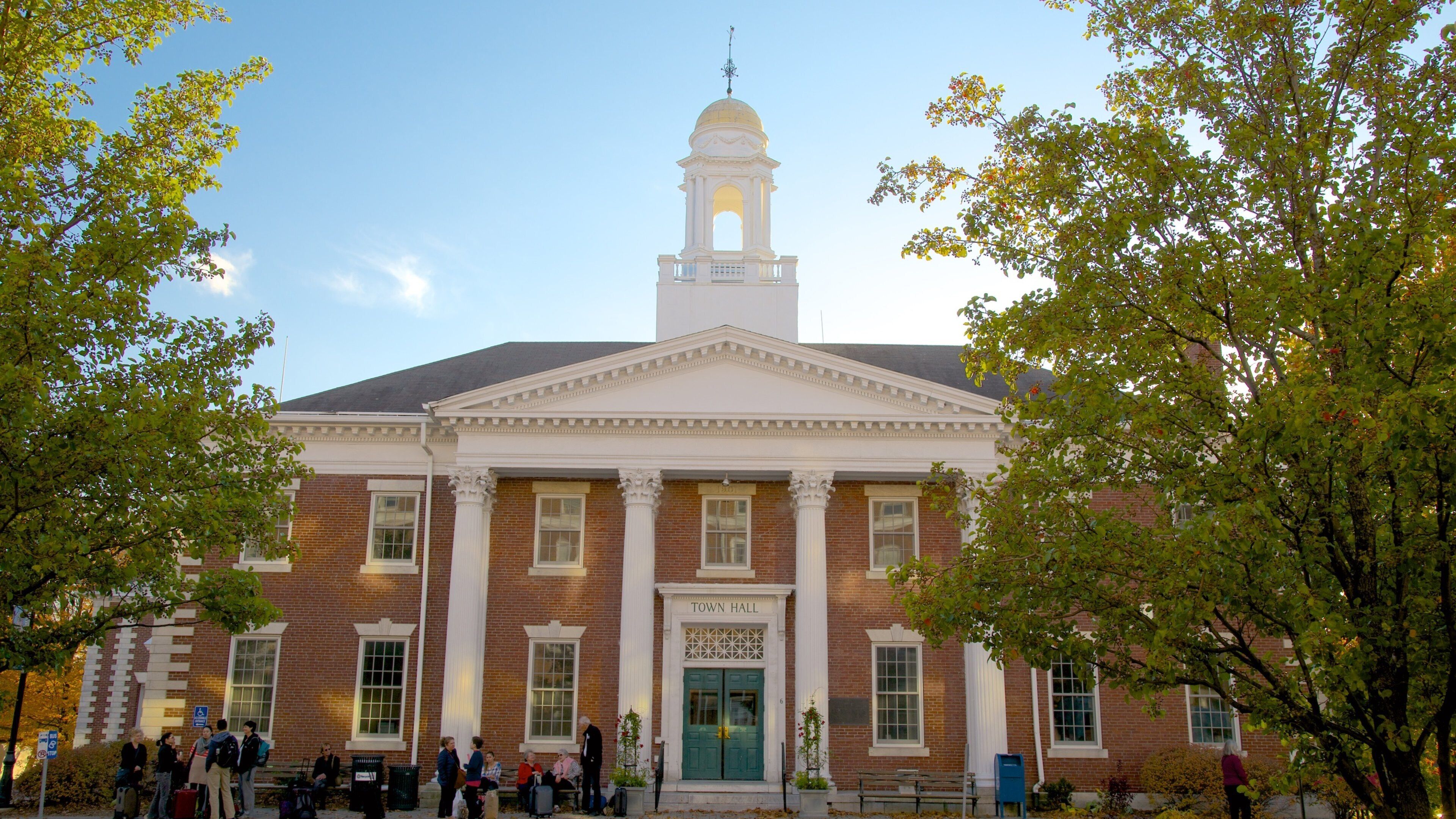 A town hall with a central dome and columns, surrounded by trees and people.