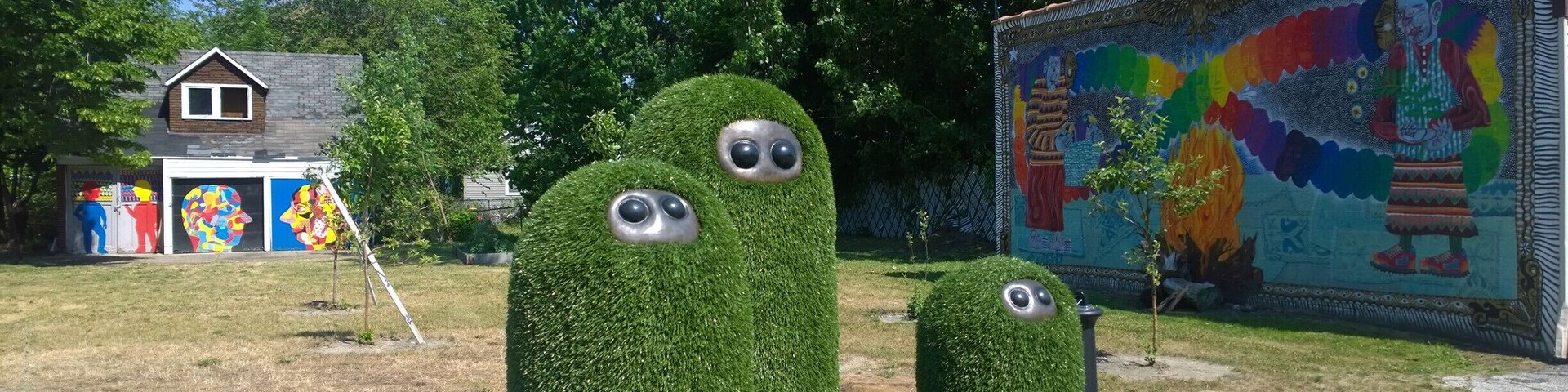 A garden with three tall, grass-covered sculptures with eyes, a colorful mural, and a house in the background.