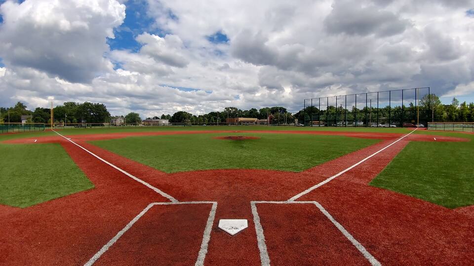 A baseball field with a red infield and green outfield, surrounded by trees and a cloudy sky.