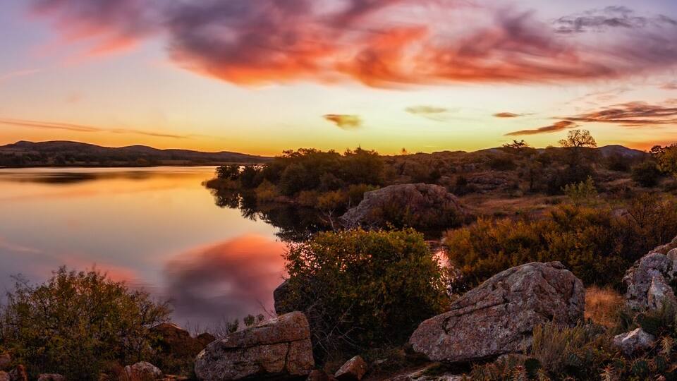 A calm lake surrounded by rocky terrain and sparse vegetation during sunset.