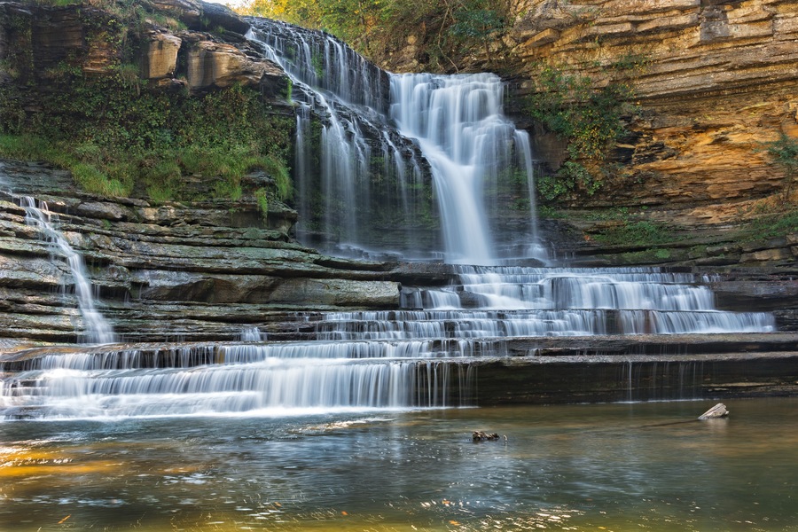 Ein mehrstufiger Wasserfall stürzt über steinige Klippen in ein ruhiges Becken.