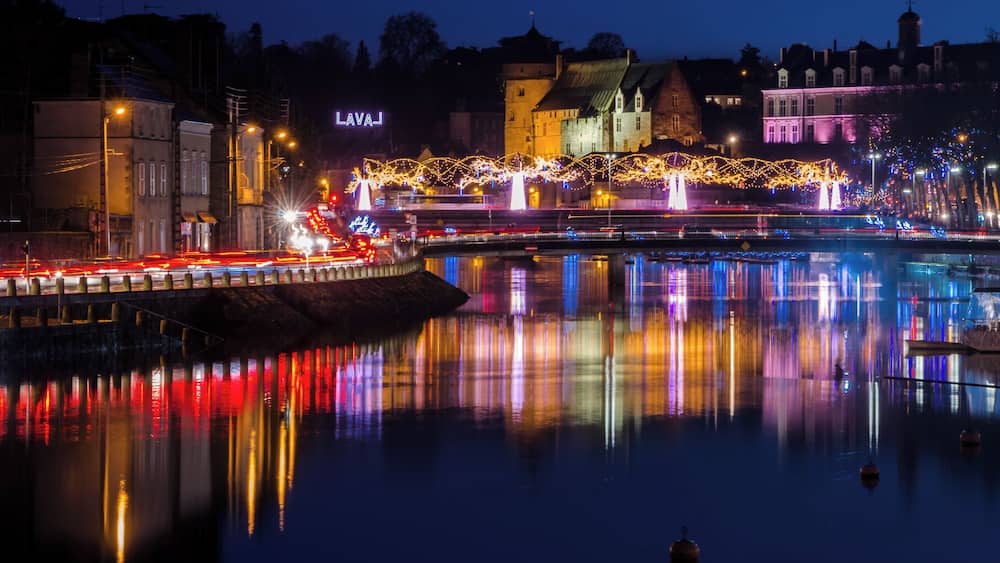 Ein beleuchtetes Stadtbild bei Nacht mit Gebäuden, einer Brücke und einem Fluss, der Lichter spiegelt.