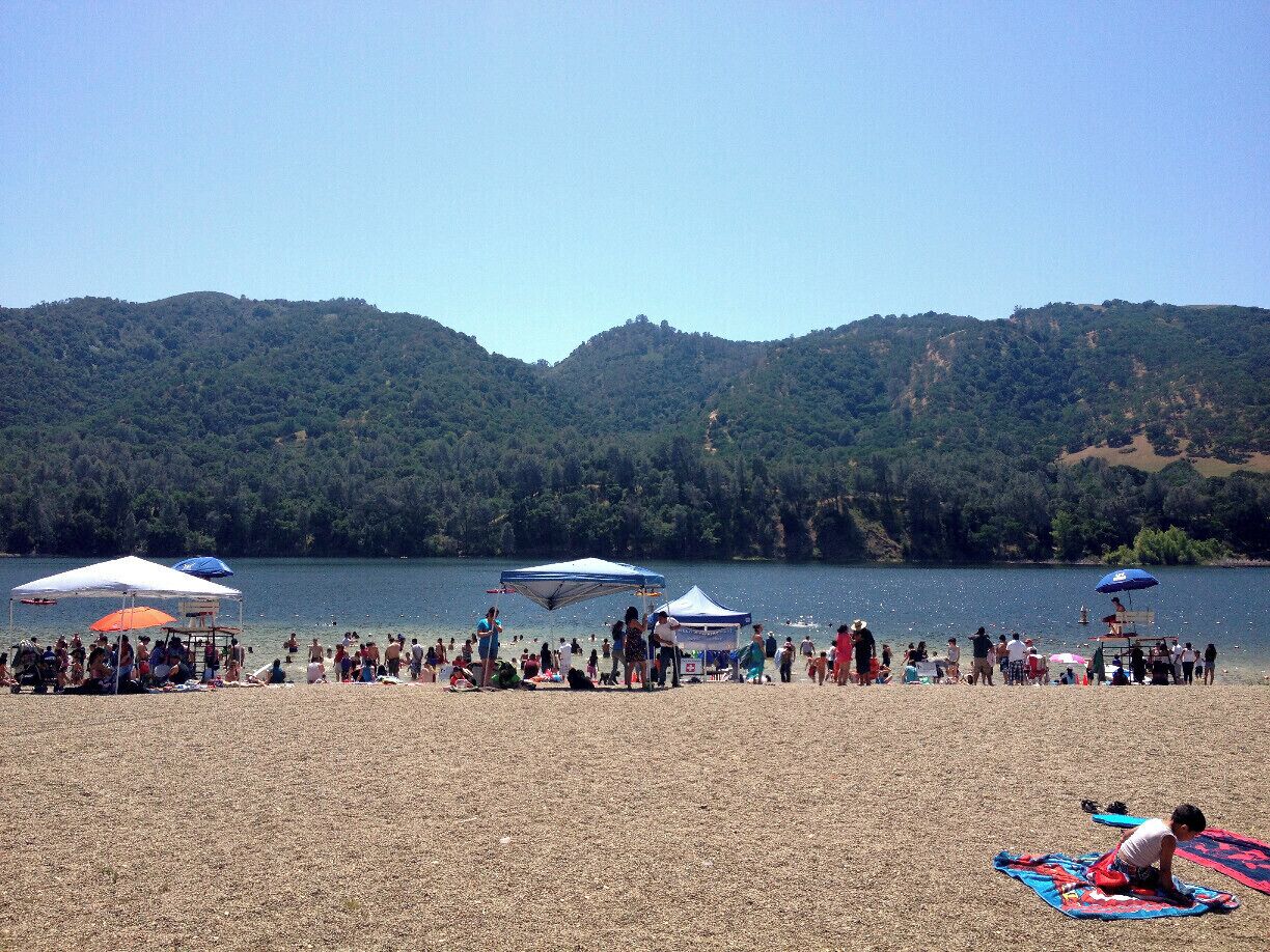 A crowded beach with people, tents, and a lake surrounded by hills.