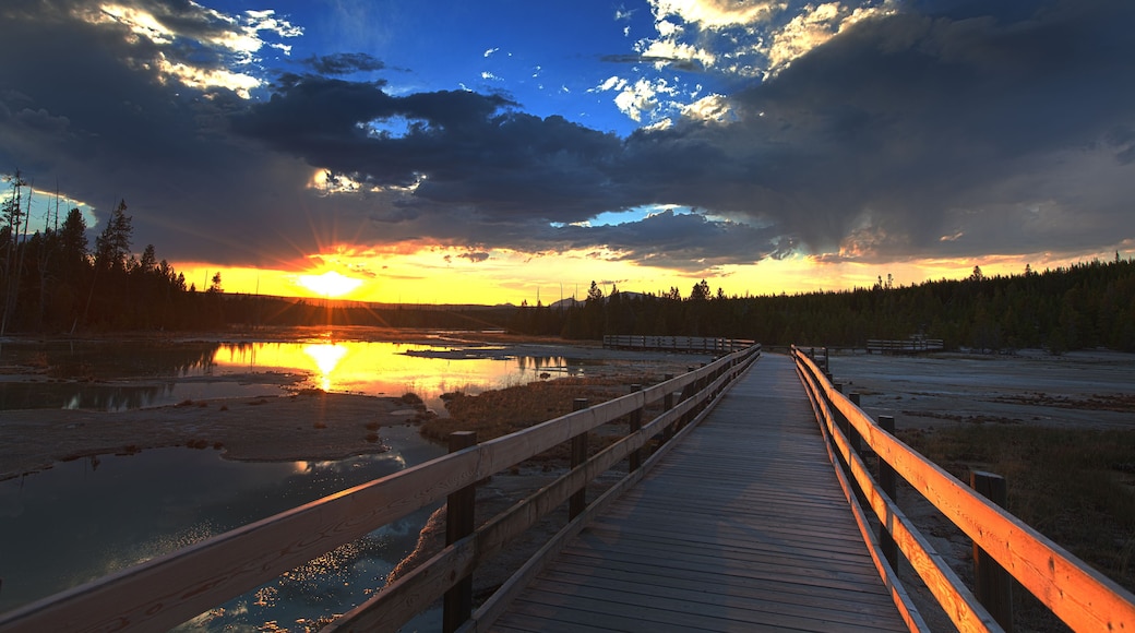 A sunset over a lake with a wooden boardwalk leading to a forested area.