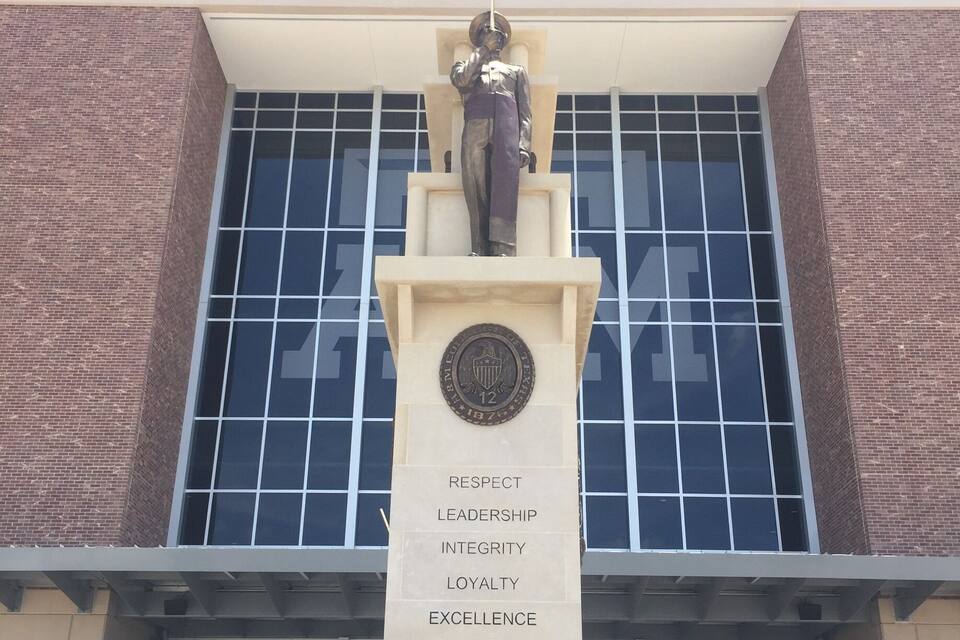 A statue with a plaque in front of a building with large windows and a sign that reads "KYLE FIELD".