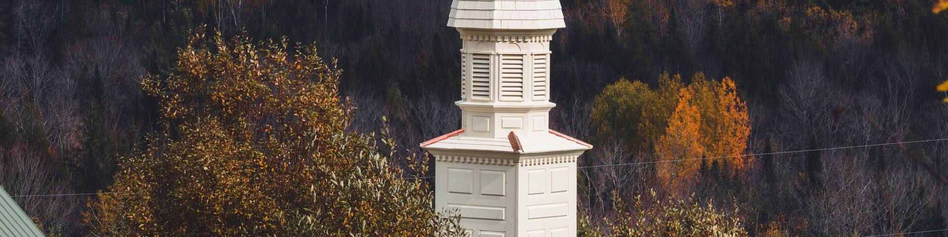 Un campanario de iglesia blanco con una estatua dorada de un ángel en la cima, rodeado de árboles de colores otoñales.