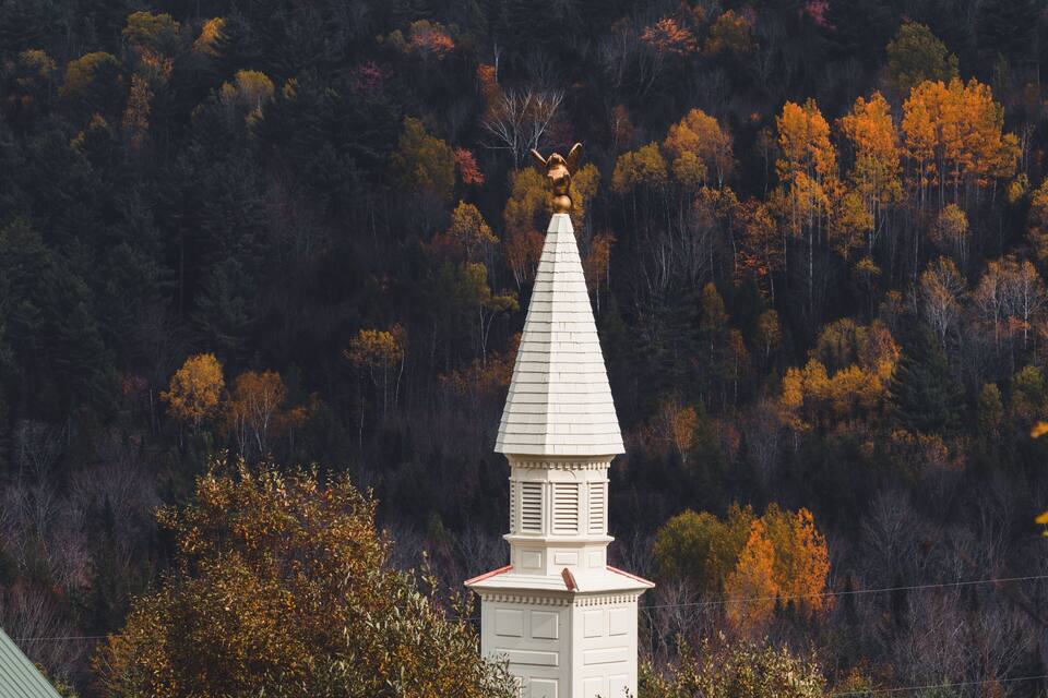 A white church steeple with a golden angel statue on top, surrounded by autumn-colored trees.
