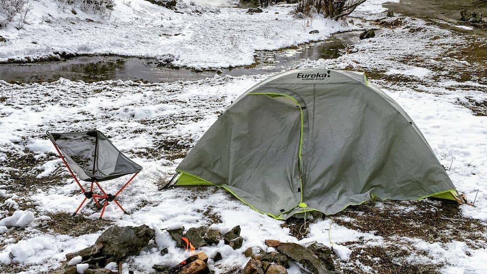 Une tente est installée près d’un ruisseau enneigé, avec un foyer à feu de camp en premier plan.