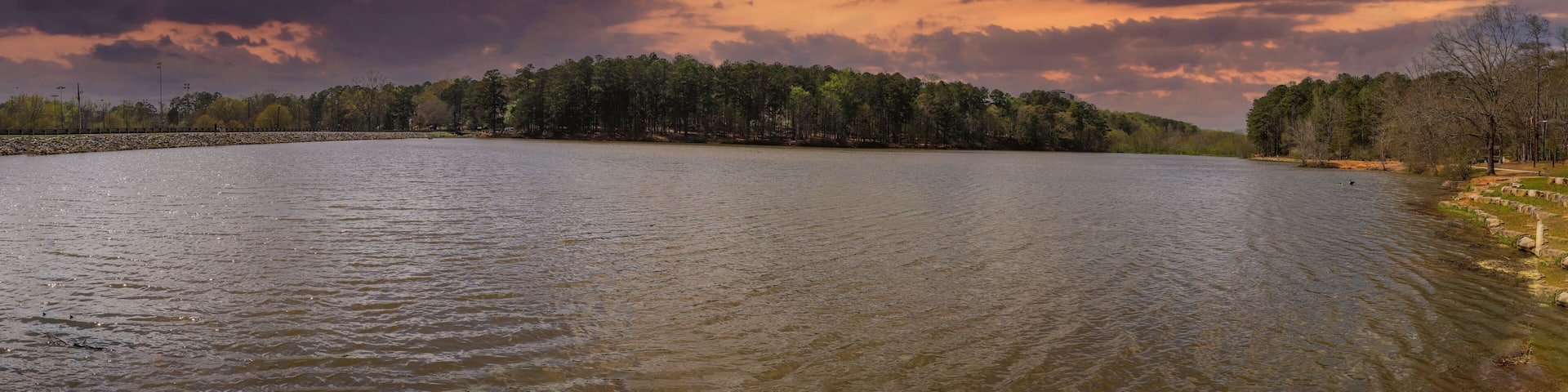 a panoramic shot of a vast rippling lake surrounded by lush green trees with powerful red clouds at sunset at Murphey Candler Park in Atlanta Georgia USA