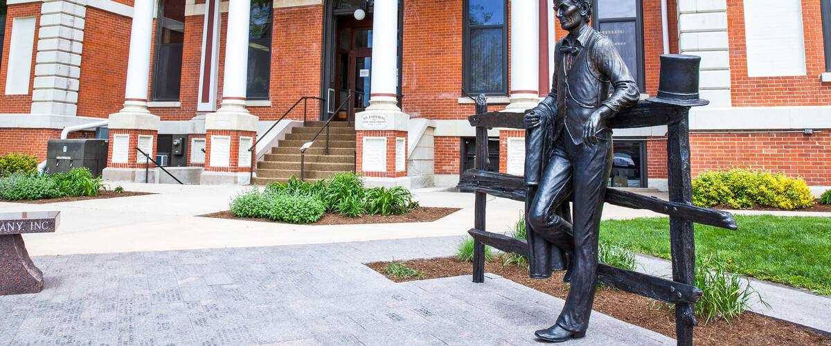A statue of a man in period clothing on a bench in front of a red brick building with white columns and a staircase.
