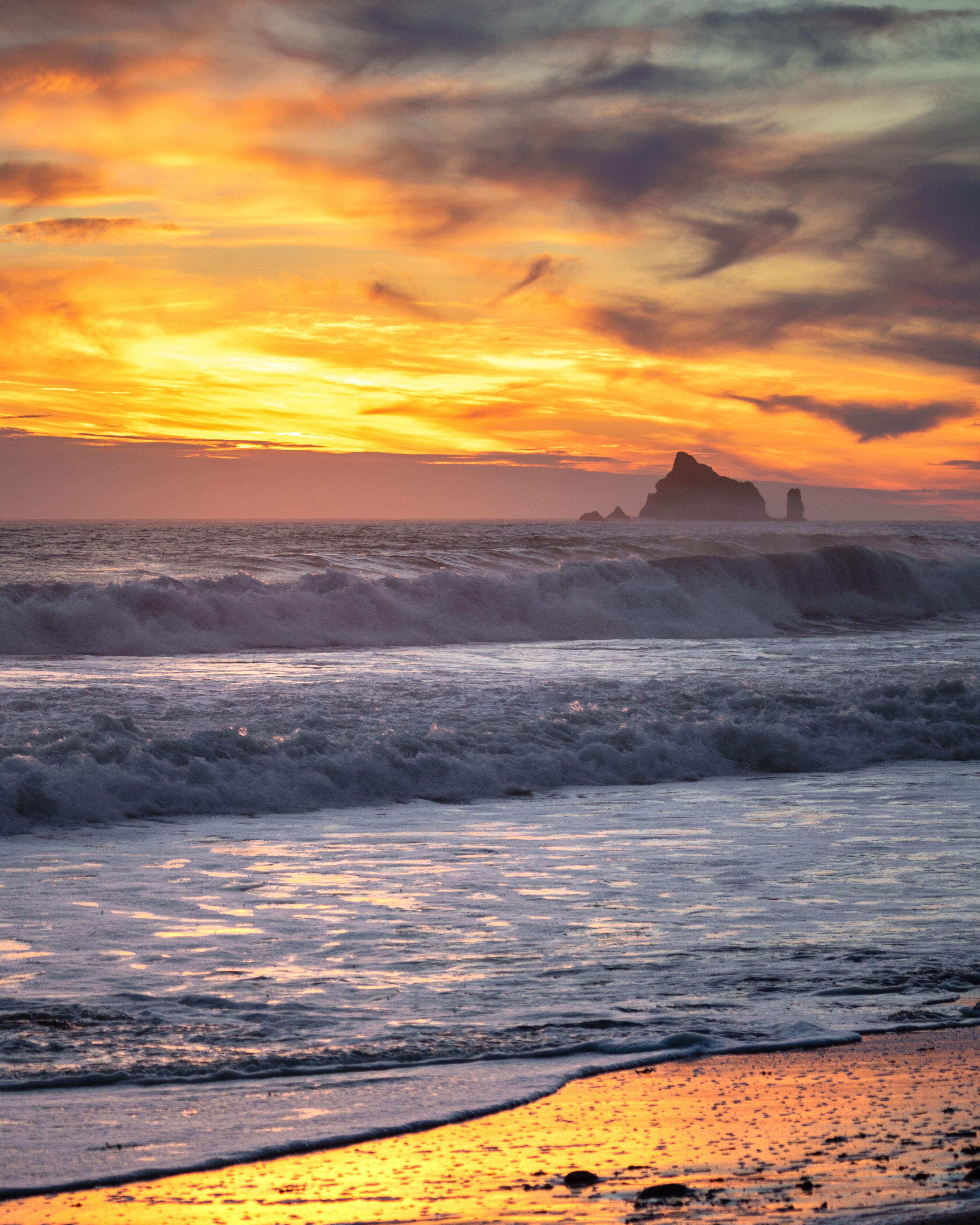 A sunset over the ocean with waves and distant rock formations.