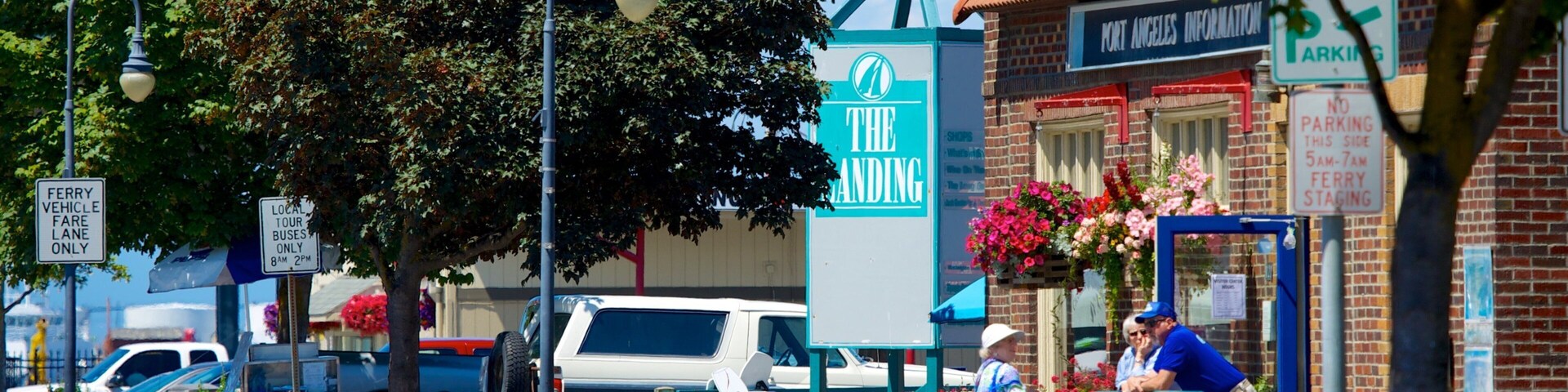 Escena callejera con una camioneta blanca, un camión azul y un coche blanco estacionados al borde de la acera. Se observan letreros que indican 'The Landing' y 'Solo vehículos para el ferry. Tarifa'. Un edificio azul muestra un cartel con la inscripción 'Port Angeles Information'.