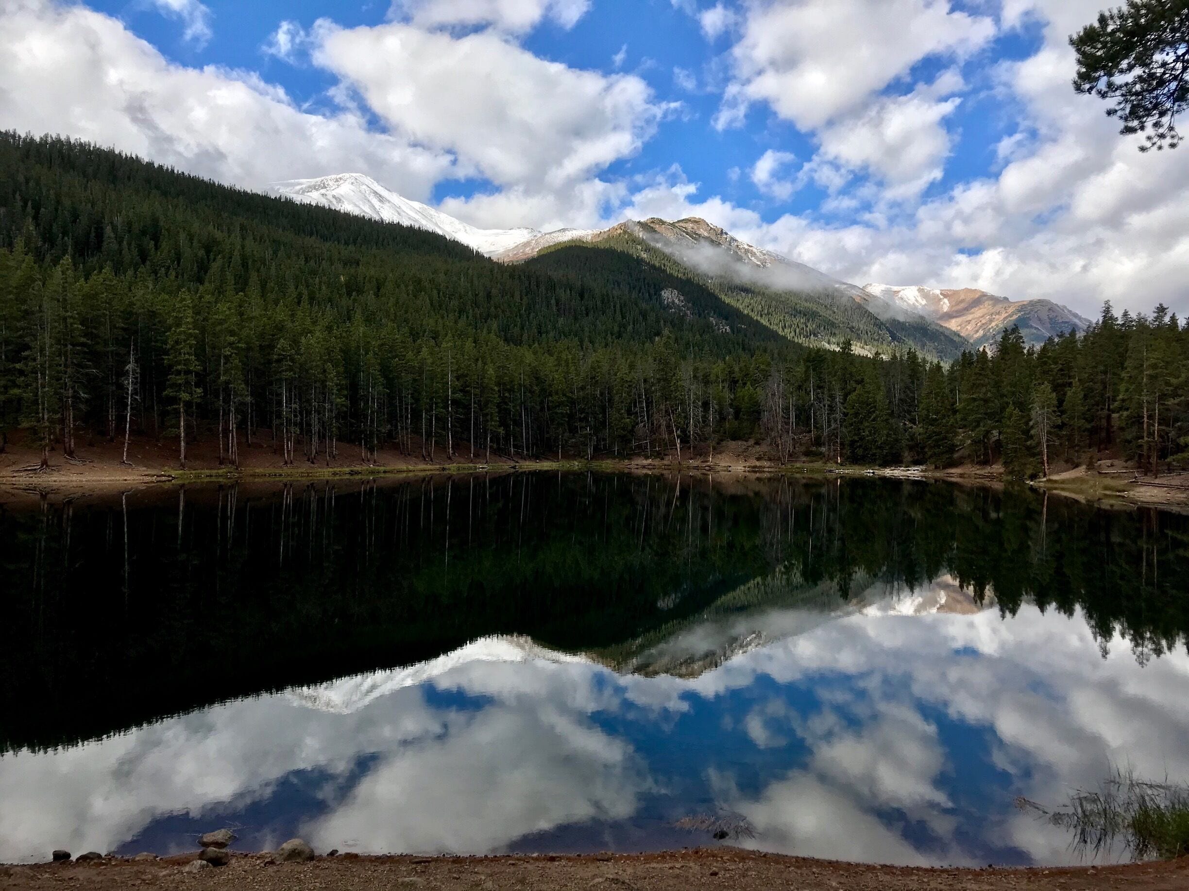A mountain range with snow-capped peaks, a dense forest, and a calm lake reflecting the sky and clouds.
