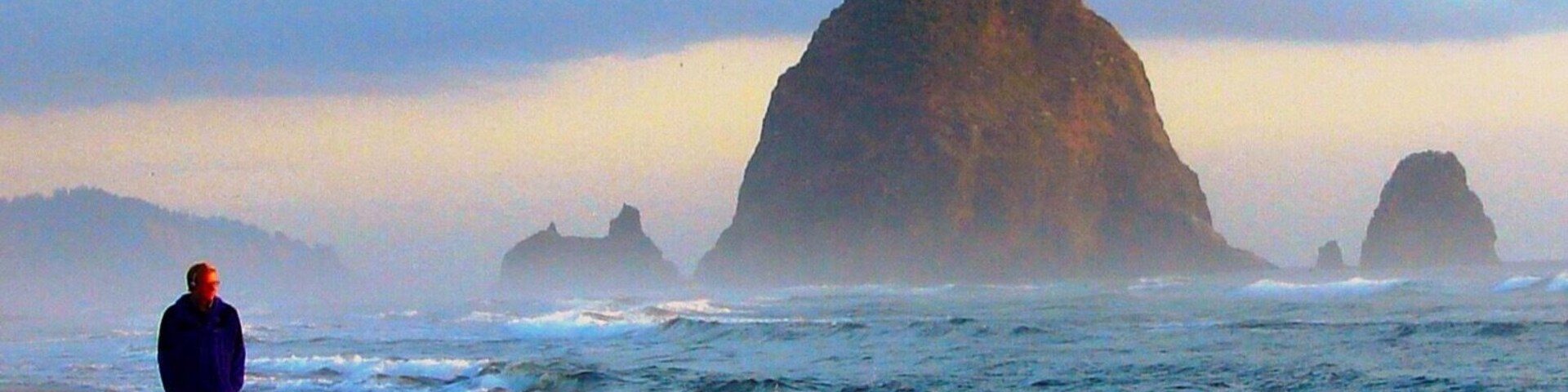 Uma pessoa caminhando numa praia, com ondas a quebrarem na costa e grandes formações rochosas ao longe.
