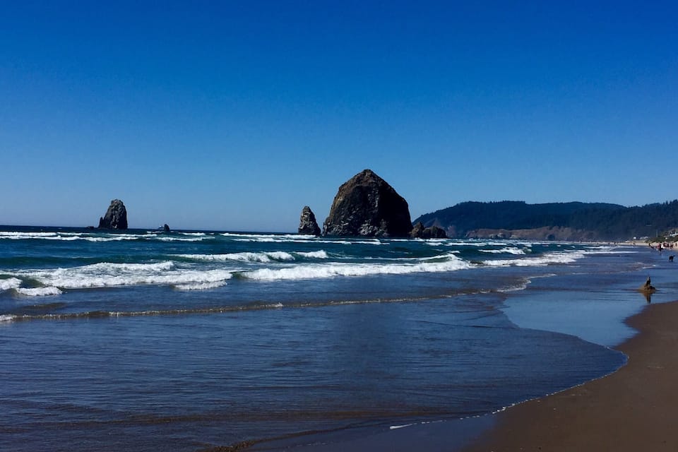 A beach with waves, a large rock formation, and a forested hillside.