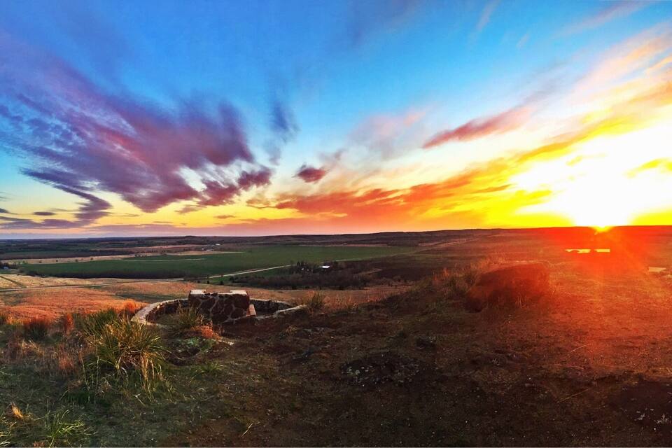 Un coucher de soleil sur un paysage vallonné, avec des champs et une construction en pierre.