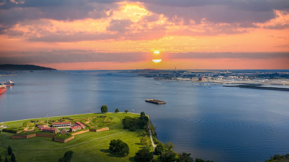 A coastal cityscape with a sunset over the water, ships, and a historic fort.