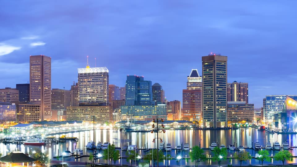 A city skyline at dusk with illuminated buildings and a river with boats.