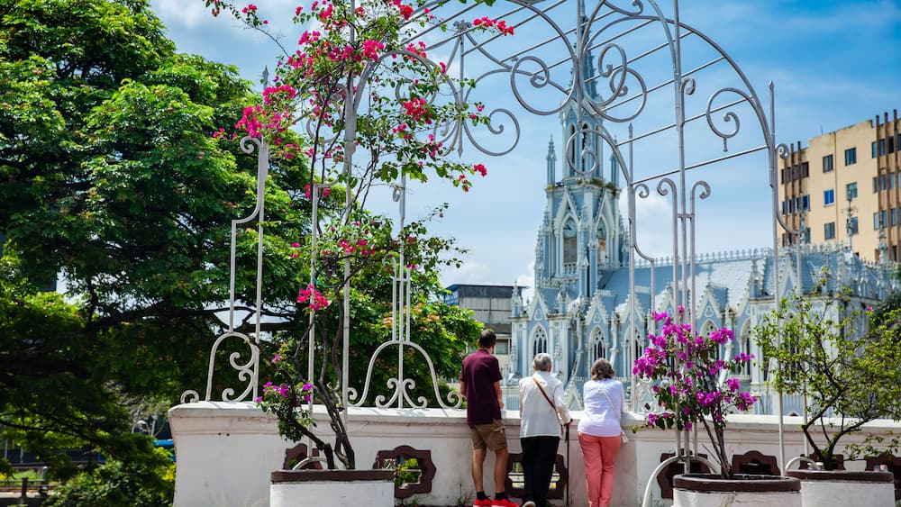 Ein schmiedeeisernes Tor mit rosa Bougainvilleen, ein Dom mit blau-weißer Architektur und ein gepflasterter Weg.