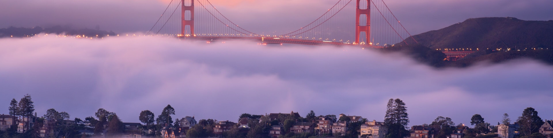The Golden Gate Bridge over fog-covered hills with a sunset sky.