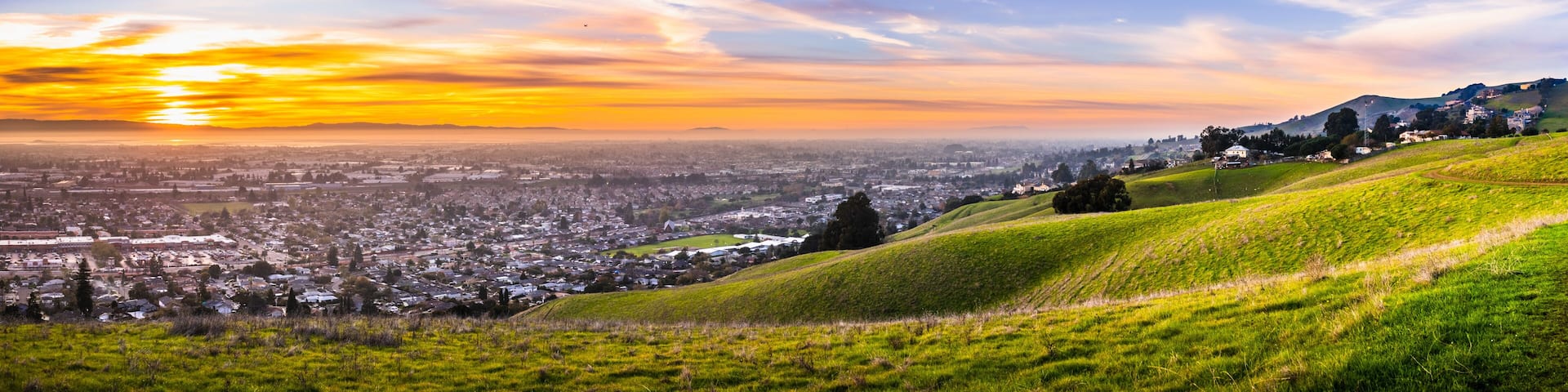 Sunset view of residential and industrial areas in East San Francisco Bay Area; green hills visible in the foreground; Hayward, California
