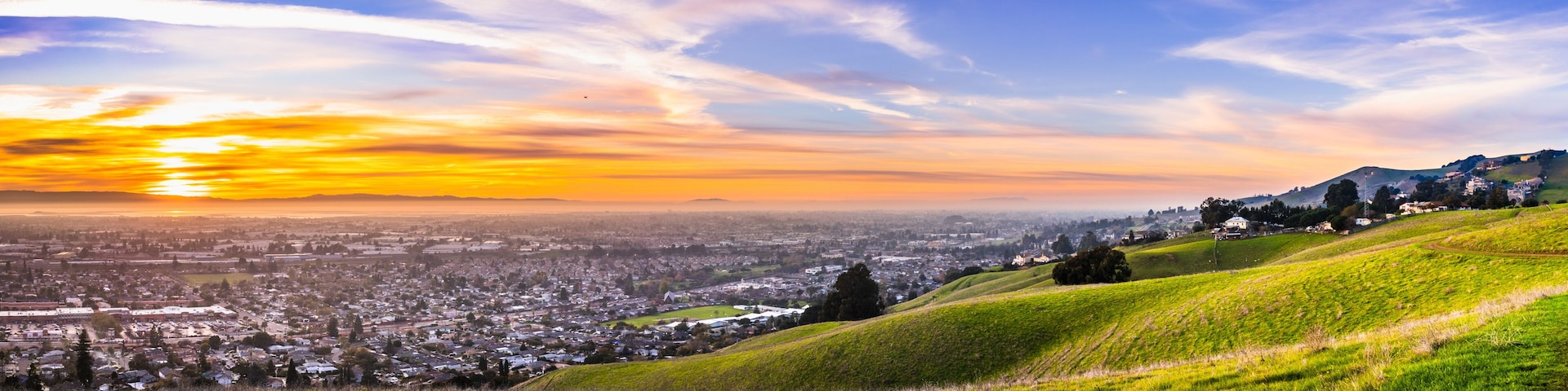 A panoramic view of a city at sunset with rolling hills in the foreground and a clear sky.