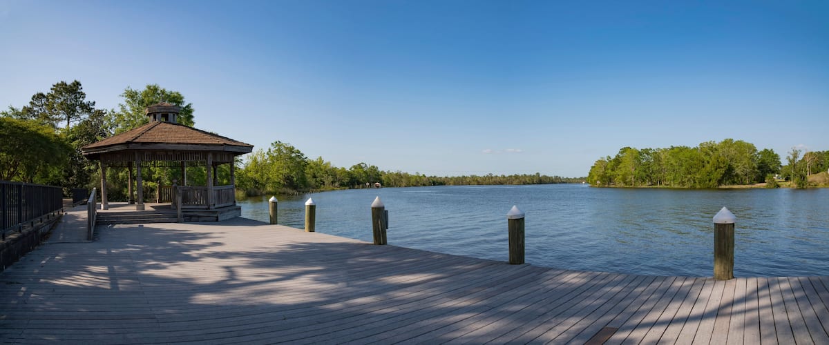 A wooden pier with a gazebo and bollards, leading to a calm lake surrounded by trees.