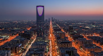 Kaupunkikuva hämärän aikaan, jossa näkyvästi esiintyy korkea pilvenpiirtäjä, jonka kaarimainen osa on valaistu violetilla valolla.