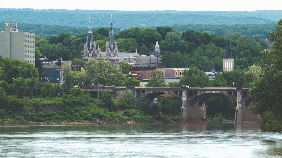 A river with a stone bridge, a church with spires, and a hill with trees in the background.