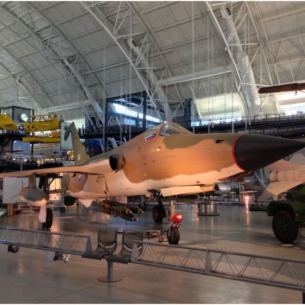 A military aircraft on display in a hangar with other aircraft and a yellow helicopter in the background.