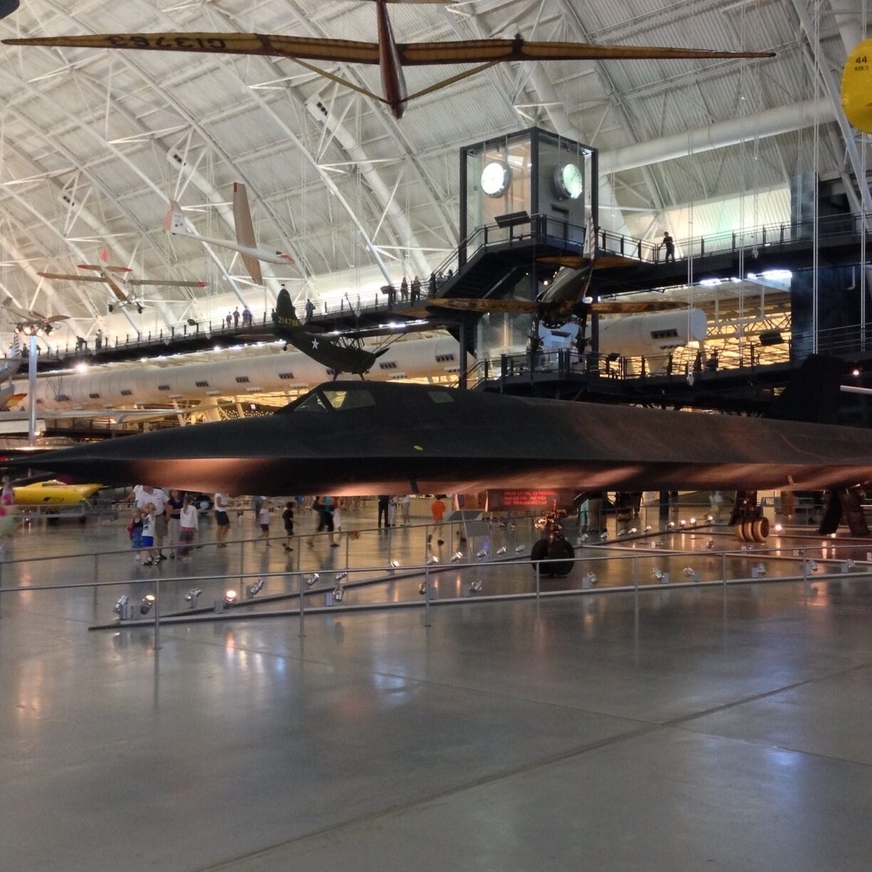 A large indoor hangar with a military aircraft on display, a clock tower, and a mezzanine level with visitors.