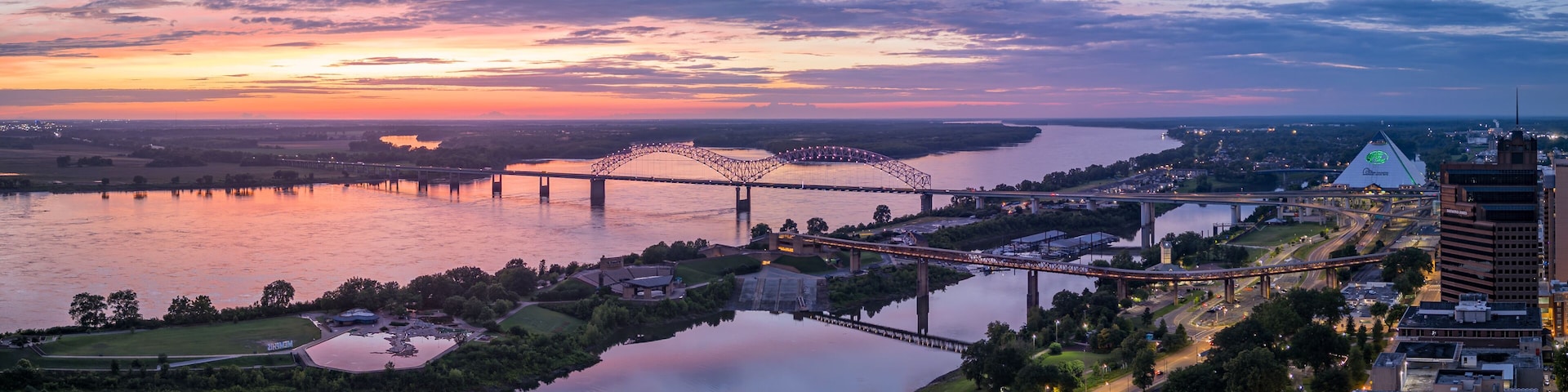 Pôr do sol sobre um rio com pontes, um cais de barcos e edifícios urbanos.