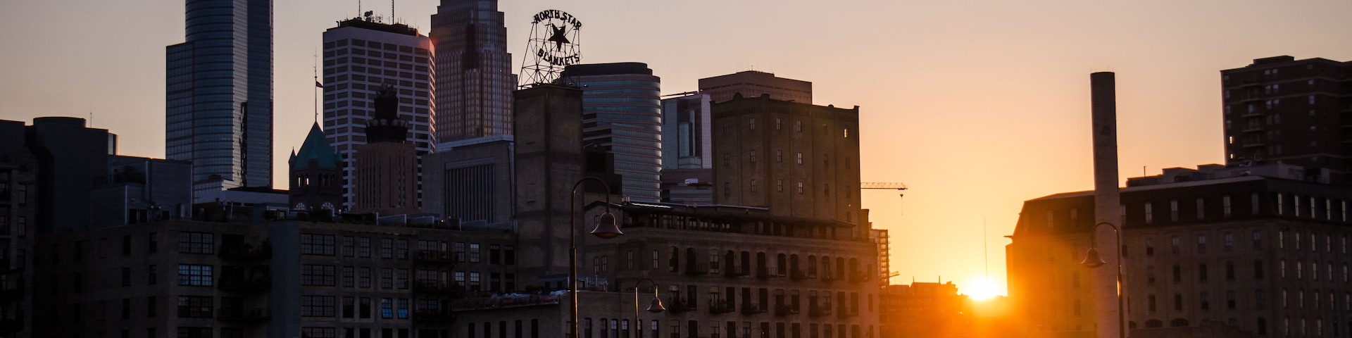 Un atardecer sobre el horizonte de una ciudad con edificios modernos e históricos, un puente y un grupo de personas en la acera.