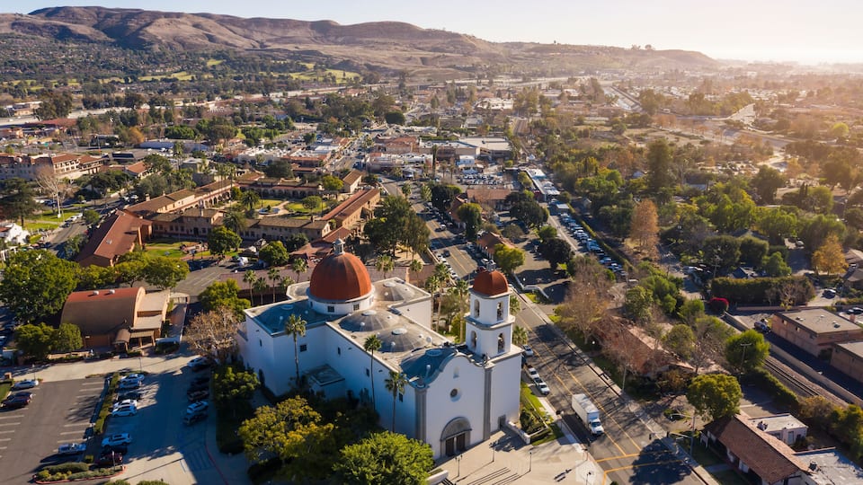 An aerial view of a town with a prominent church featuring two domes, surrounded by residential areas and a hilly landscape in the background.