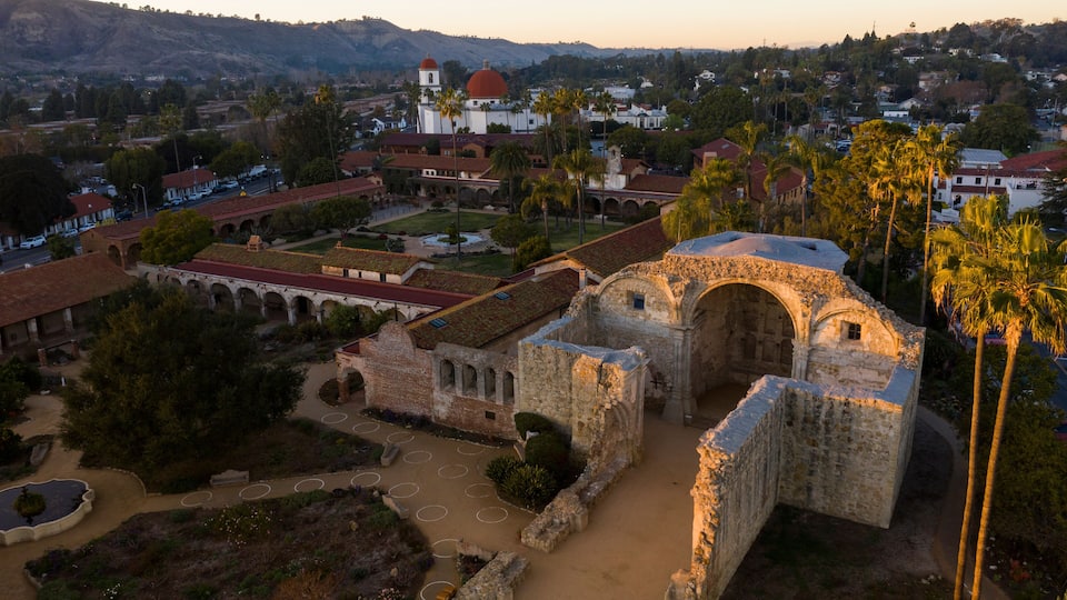 An ancient stone structure with arches and a dome, surrounded by palm trees and a town in the background.