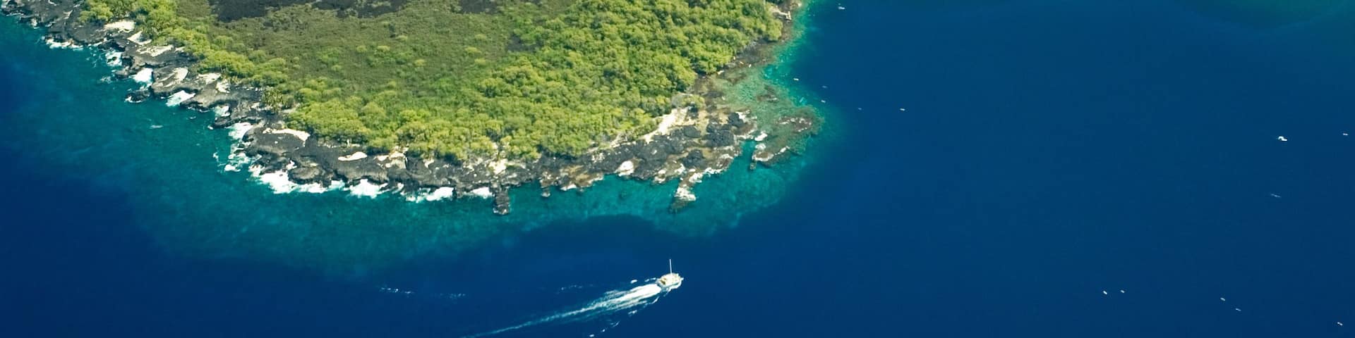 An aerial view of a coastal area with lush green hills, a clear blue sea, and a boat creating a wake.