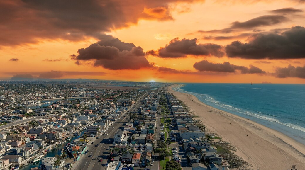 an aerial shot of the vast miles of homes and lush green trees around Huntington Harbour with blue water, majestic mountain ranges, cars driving on the street and powerful clouds at sunset