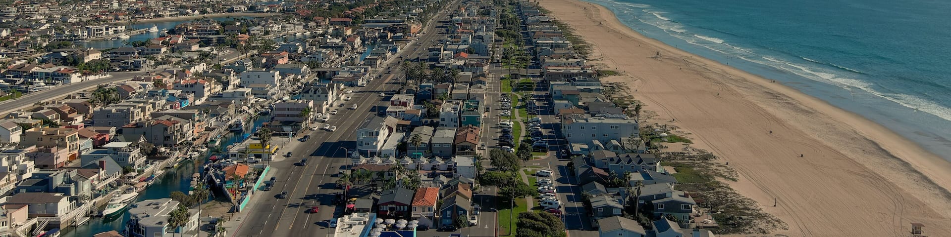 an aerial shot of the vast miles of homes and lush green trees around Huntington Harbour with blue water, majestic mountain ranges, cars driving on the street and powerful clouds at sunset