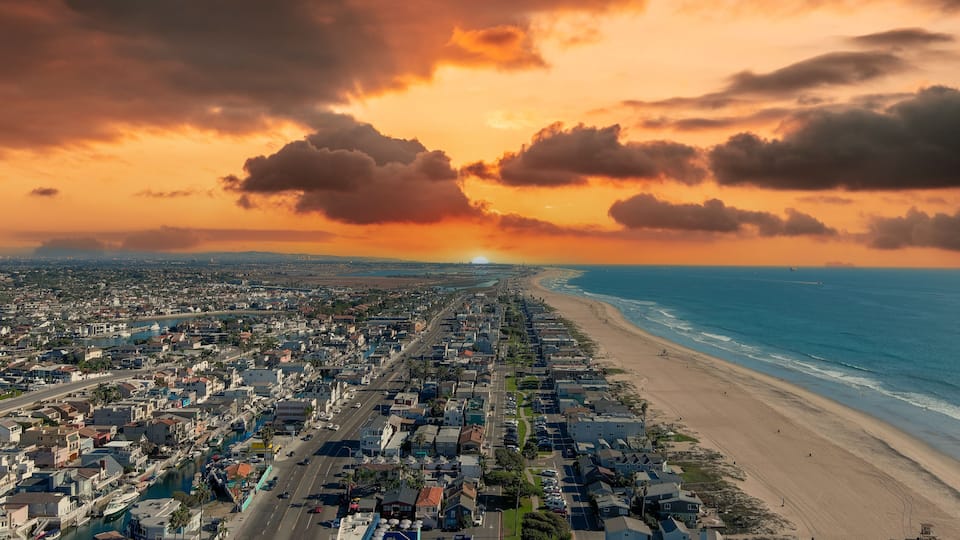 an aerial shot of the vast miles of homes and lush green trees around Huntington Harbour with blue water, majestic mountain ranges, cars driving on the street and powerful clouds at sunset