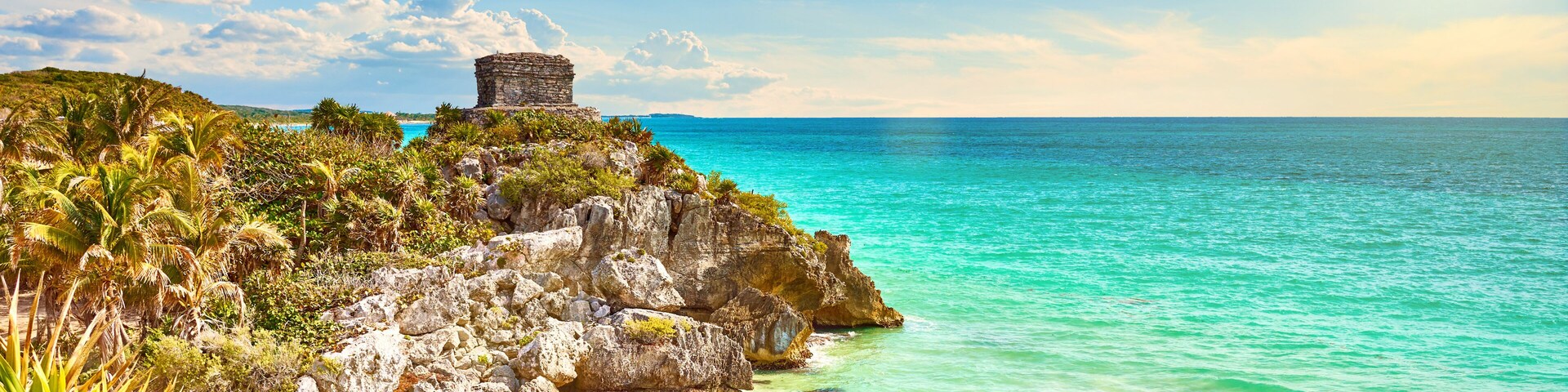 Un paisaje costero con un mar turquesa transparente, una playa arenosa, afloramientos rocosos y una pequeña estructura de piedra en una colina.