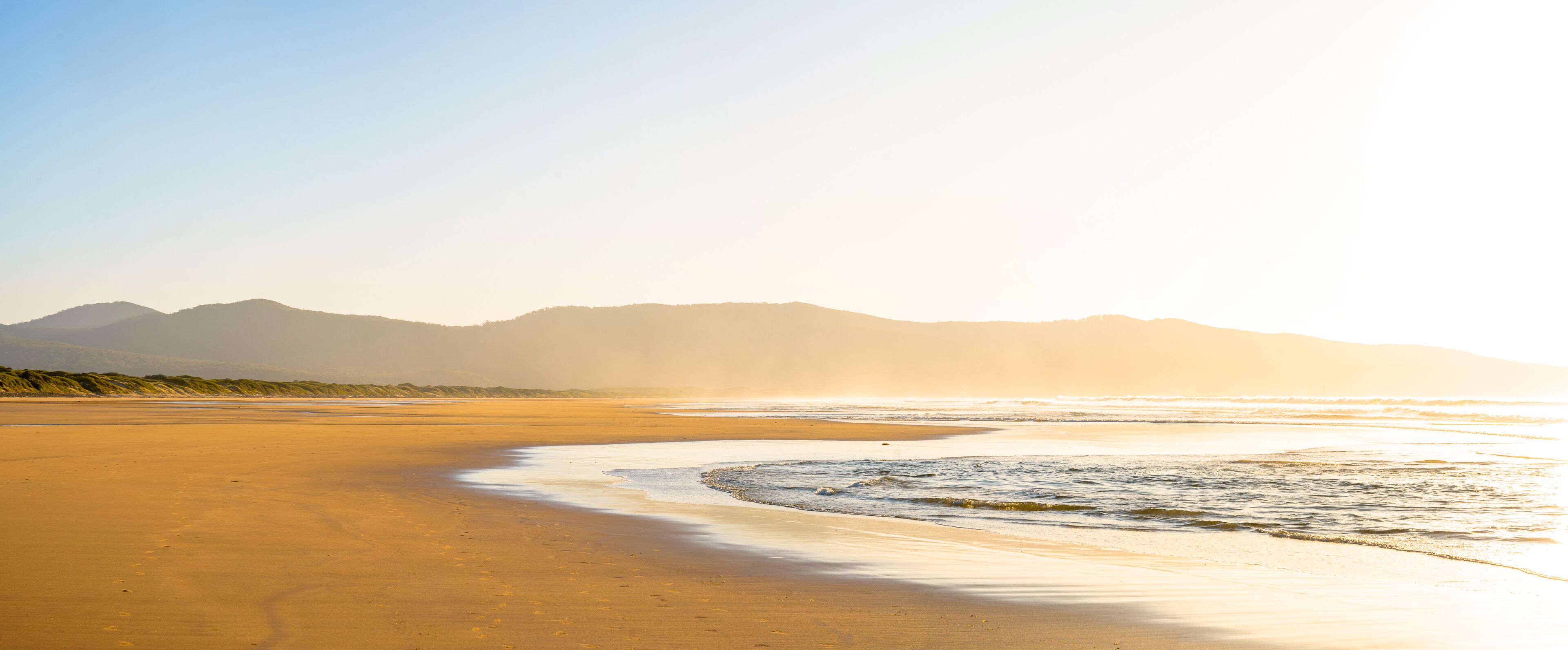 A beach with a curved shoreline, waves, and distant mountains under a clear sky.