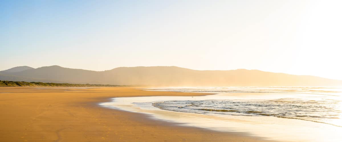 A beach with a curved shoreline, waves, and distant mountains under a clear sky.