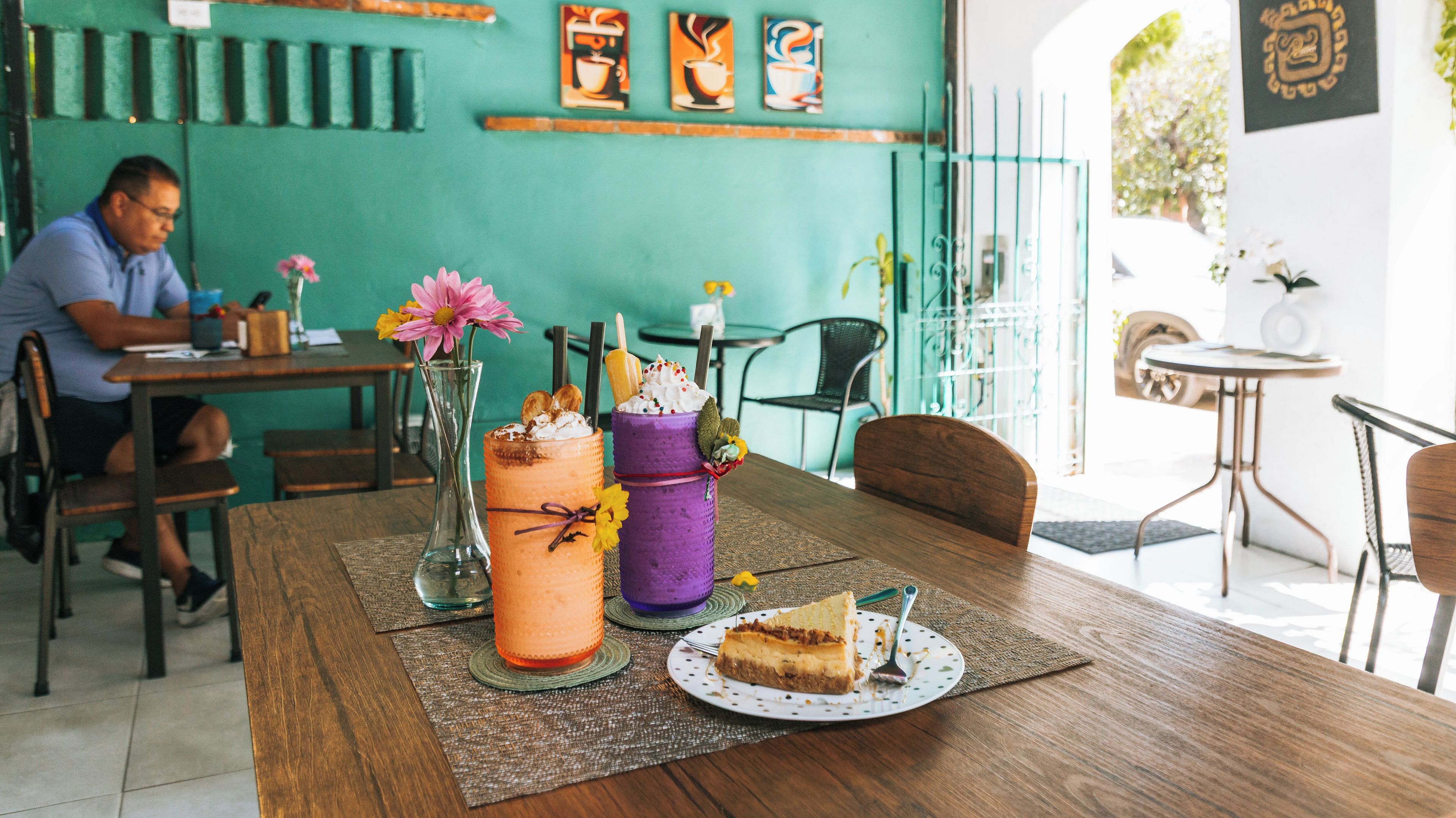 A cafe with a wooden table, two colorful desserts, a vase with flowers, and a man sitting at a table.