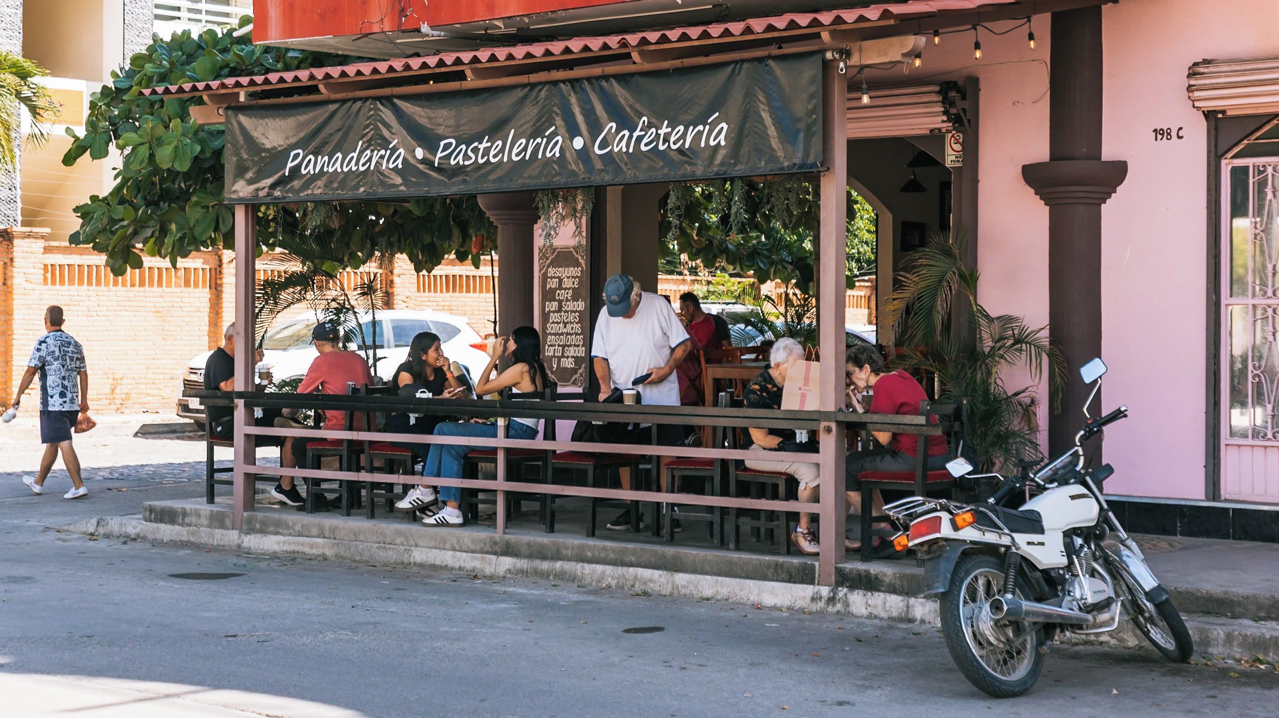 A street-side cafe named 'Panaderia Pastelería Cafetería' with outdoor seating and a motorcycle parked nearby.