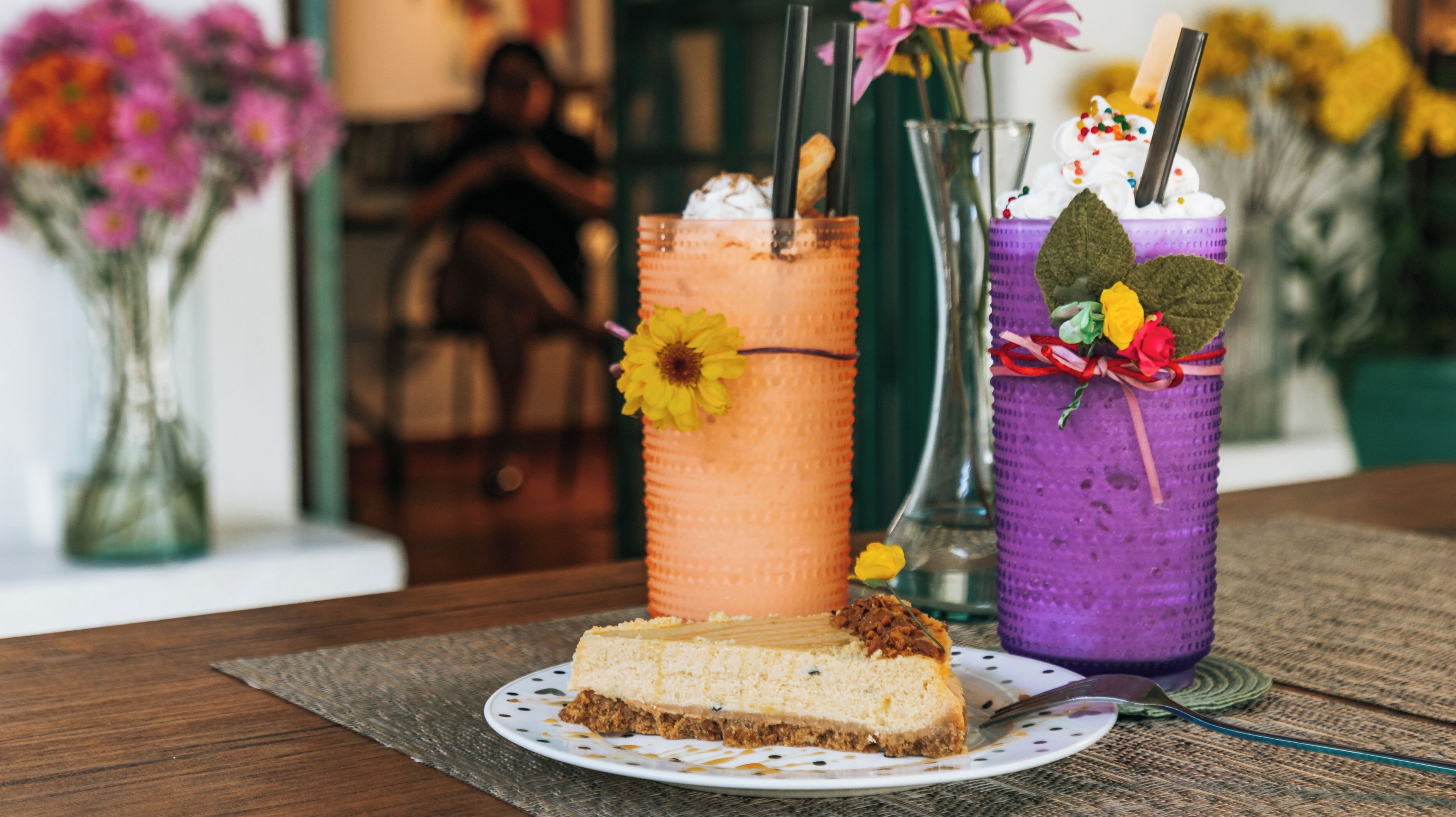A slice of cheesecake on a plate with two colorful drinks in the background.