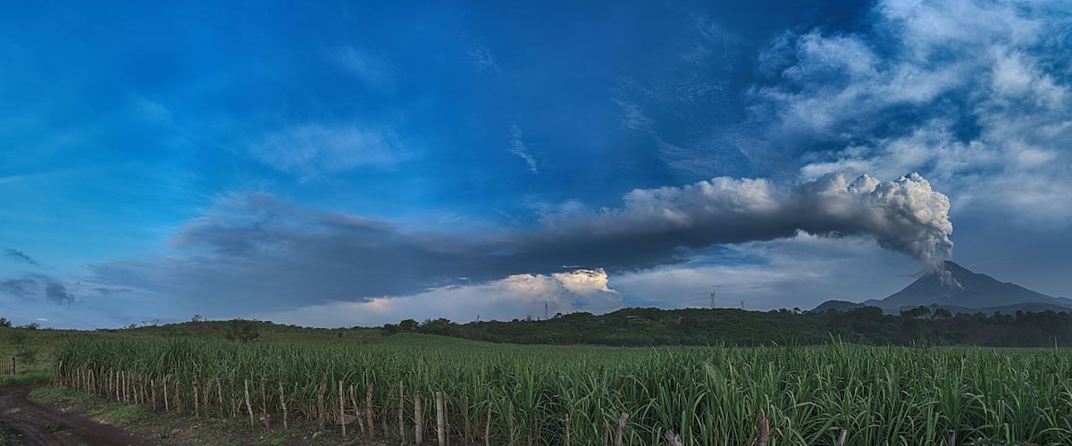 A volcanic landscape with a clear sky, a field of crops, and a distant volcano emitting smoke.